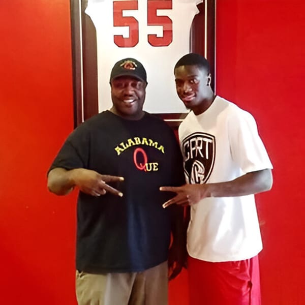 Two men, one in an Alabama Que shirt, making peace signs in front of a red wall.