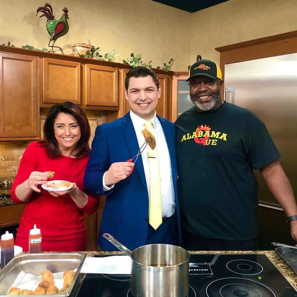 Three people, including the Alabama Que owner, smiling in a kitchen with fried food and sauces.