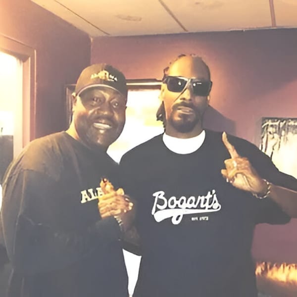 Snoop Dogg in sunglasses and a man in an Alabama Que hat pose together.