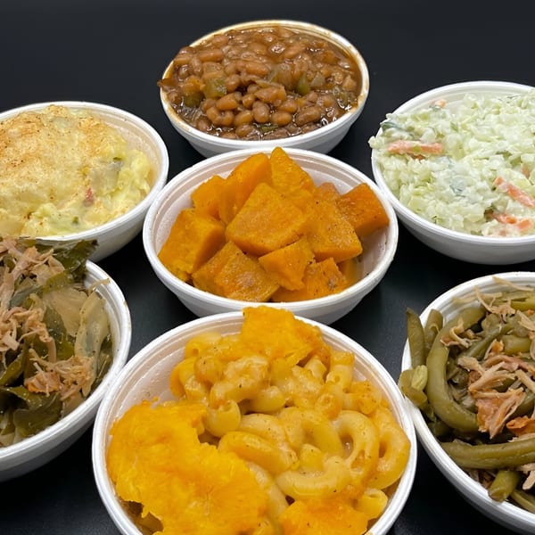 Six white bowls filled with various Alabama Que BBQ sides, including baked beans, mac and cheese, and coleslaw.
