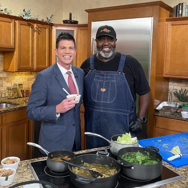 Two men, one in a suit and one in Alabama Que BBQ attire, cooking in a kitchen.