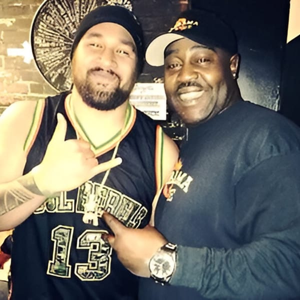 Two men smiling at the camera, one in a basketball jersey, the other in an Alabama Que hat.