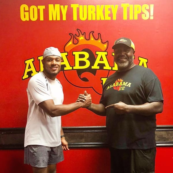 Two men shaking hands in front of a red Alabama Que BBQ wall with yellow lettering.