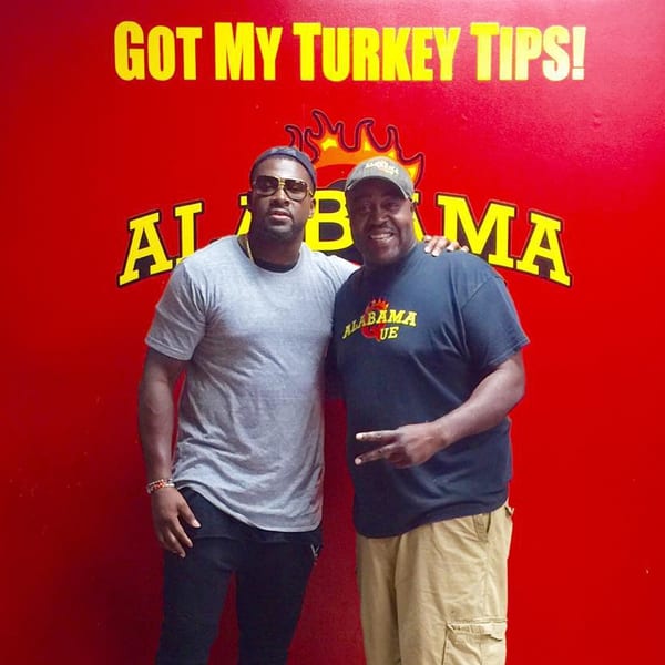 Two men posing in front of a red wall with "GOT MY TURKEY TIPS! ALABAMA QUE" in yellow text.
