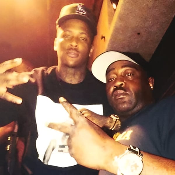 Two men in hats pose for a photo, one making a peace sign, in a dimly lit setting.