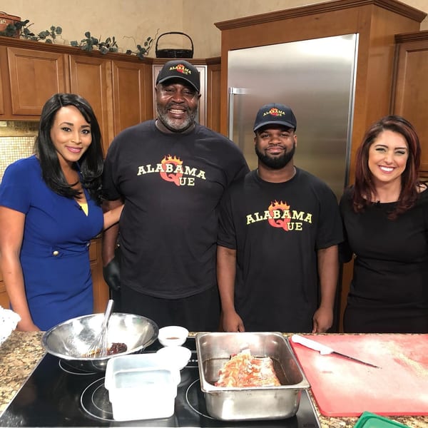 Two men in Alabama Que shirts and two women in a kitchen setting, with raw meat and ingredients.