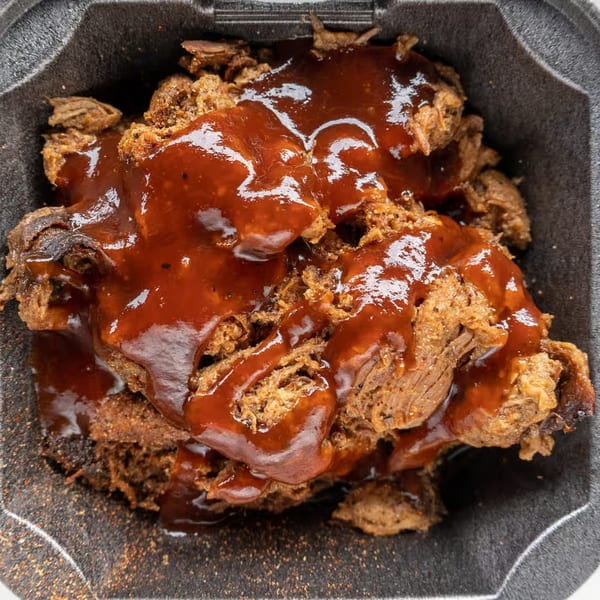 Close-up of Alabama Que's pulled pork, generously topped with glistening BBQ sauce in a black container.